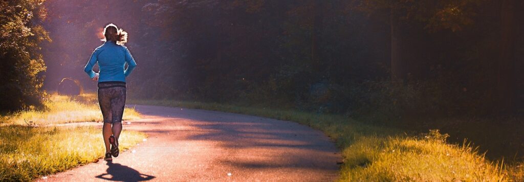 Na zdjęciu widać kobietę biegnącą lewą stroną drogi asfaltowej w lesie
