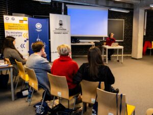 Na zdjęciu widać uczestniczki warsztatów oglądające uczestniczkę ćwiczącą wystąpienie przed mikrofonem.