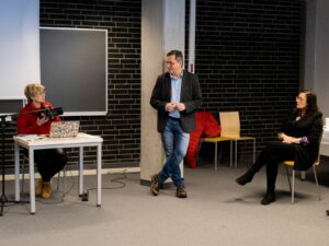 Na zdjęciu widac uczestniczkę warsztatów czytającą tekst przed mikrofonem oraz prowadzących warsztaty.