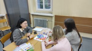 Zdjęcie przedstawia kilka osób siedzących przy jednym stole i grających w gry planszowe na tle grzejnika oraz okna