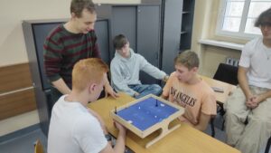 Zdjęcie przedstawia kilka osób siedzących przy jednym stole i grających w grę na drewnianym podeście wraz z niebieską górną częścią na której znajdują się elementy poruszane od spodu magnesem