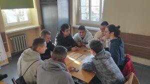 Zdjęcie przedstawia kilka osób wspólnie grających w grę planszową przy jednym stole