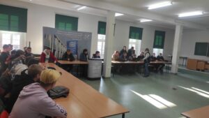 Zdjęcie przedstawia kilka osób siedzących przy stolikach i słuchających wykładu na temat technologii dronowych na tle banerów reklamowych oraz okien z zielonymi roletami
