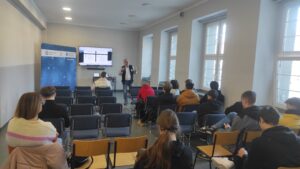 Zdjęcie przedstawia prezentera opowiadającego o technologiach AI w przemyśle na tle telewizora oraz ścianki promocyjnej Politechniki Śląskiej
