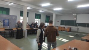 Zdjęcie przedstawia kilka osób siedzących przy stolikach i słuchających wykładu na temat technologii dronowych na tle banerów reklamowych oraz okien z zielonymi roletami, na pierwszym planie znajdują się prezenterzy