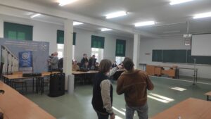 Zdjęcie przedstawia kilka osób siedzących przy stolikach i słuchających wykładu na temat technologii dronowych na tle banerów reklamowych oraz okien z zielonymi roletami, na pierwszym planie znajdują się prezenterzy