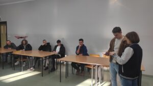 Zdjęcie przedstawia kilka osób siedzących przy stolikach i słuchających wykładu na temat technologii dronowych