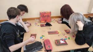 Zdjęcie przedstawia cztery osoby grające w planszówkę przy jednym stoliku