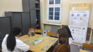 Zdjęcie przedstawia kilka osób grających w grę planszową na tle okna oraz czarnych szaf