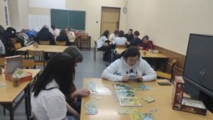 Zdjęcie przedstawia dużą liczbę osób grających w planszówki w sali lekcyjnej na tle tablicy