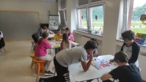 Zdjęcie przedstawia dużą ilość dzieci grających w planszówki przy kilku stołach w sali lekcyjnej na tle okien oraz tablic do pisania