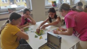 Zdjęcie przedstawia cztery osoby grające w planszówkę na zielonej planszy, obok nich znajduje się otwarte pudełko od planszówki