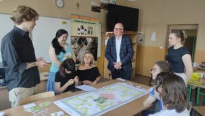 Zdjęcie przedstawia kilka osób siedzących przy dużym stole oraz kilka osób stojących obok niego, na którym znajduje się ogromna plansza gry oraz duża ilość elementów gry w postaci kart oraz instrukcji.