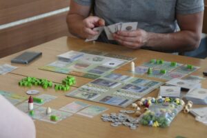 Zdjęcie przedstawia mężczyznę siedzącego przy stole na którym znajdują się karty oraz figurki do gry planszowej, trzymającego karty do gry