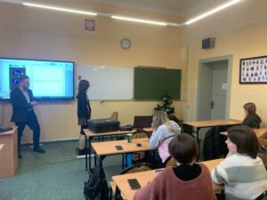 Zdjęcie przedstawia prezentera pokazującego slajdy z informacjami oraz uczestników warsztatów którzy siedzą przy ławkach klasowych