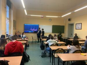 Zdjęcie przedstawia prezentera pokazującego wykres oraz uczestników warsztatów którzy siedzą przy ławkach klasowych