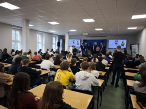 Sala wypełniona jest uczniami słuchającymi osób na scenie. Kliknięcie powoduje zwiększenie zdjęcia.