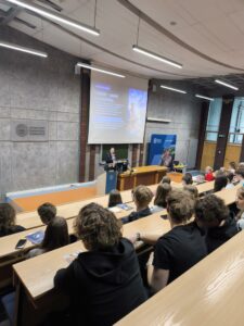 Katarzyna Dohn wygłasza prezentację na podeście. Uczestnicy spotkania siedzą w ławkach i słuchają. Kliknięcie powoduje zwiększenie zdjęcia.