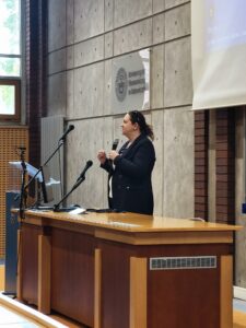 zbliżenie na Katarzynę Dohn, wygłaszającą prezentację na podeście. Kliknięcie powoduje zwiększenie zdjęcia.