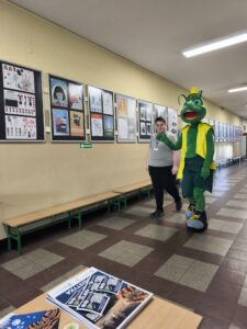 Maskotka szkolna i uczeń idą szkolnym korytarzem. Kliknięcie powoduje zwiększenie zdjęcia.