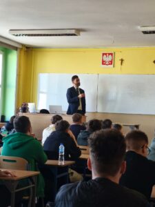 Dzieci siedzą w szkolnych ławkach i słuchają osoby prezentującej. Kliknięcie powoduje zwiększenie zdjęcia.