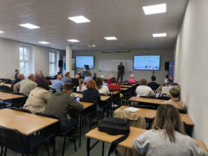 Uczestnicy spotkania siedzą w ławkach na auli i słuchają wygłaszanej prezentacji. Kliknięcie powoduje zwiększenie zdjęcia.