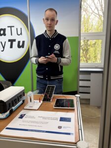Przy stanowisku stoi pracownik Politechniki. Na blacie znajduje się plakat informacyjny projektu COZEiTW oraz wiatraczek podłączony do paneli na światło. Kliknięcie powoduje zwiększenie zdjęcia.