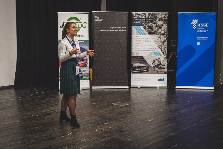 Na zdjęciu doktorantka opowiadająca o pracy naukowej, w tle bannery sponsorów konkursu