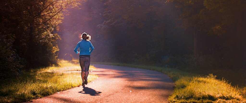 Na zdjęciu widać kobietę biegnącą lewą stroną drogi asfaltowej w lesie