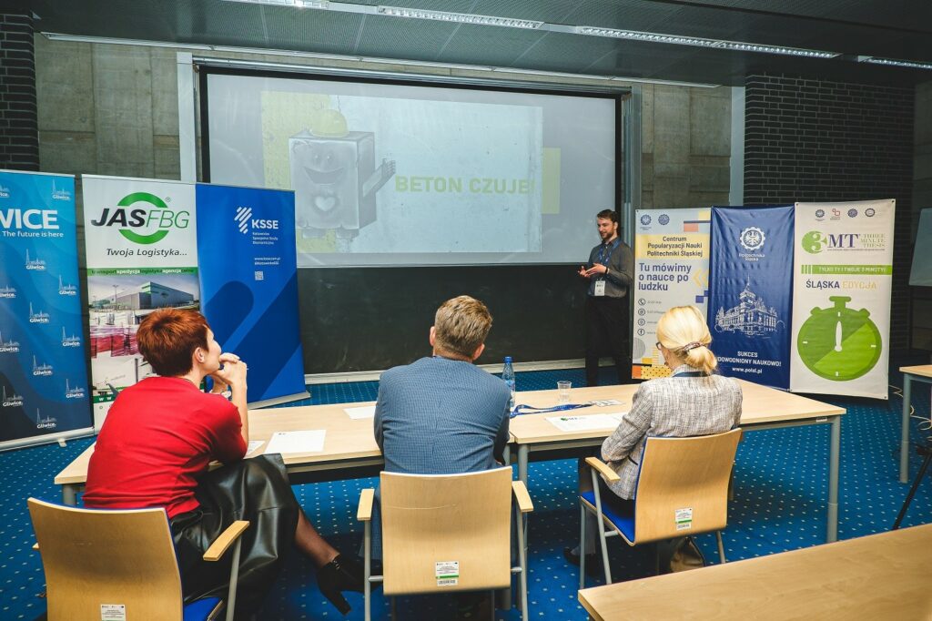 Na zdjęciu widać uczestnika konkursu Three Minute Thesis przedstawiającego prezentacje. Na pierwszym planie znajdują się trzy osoby jury,