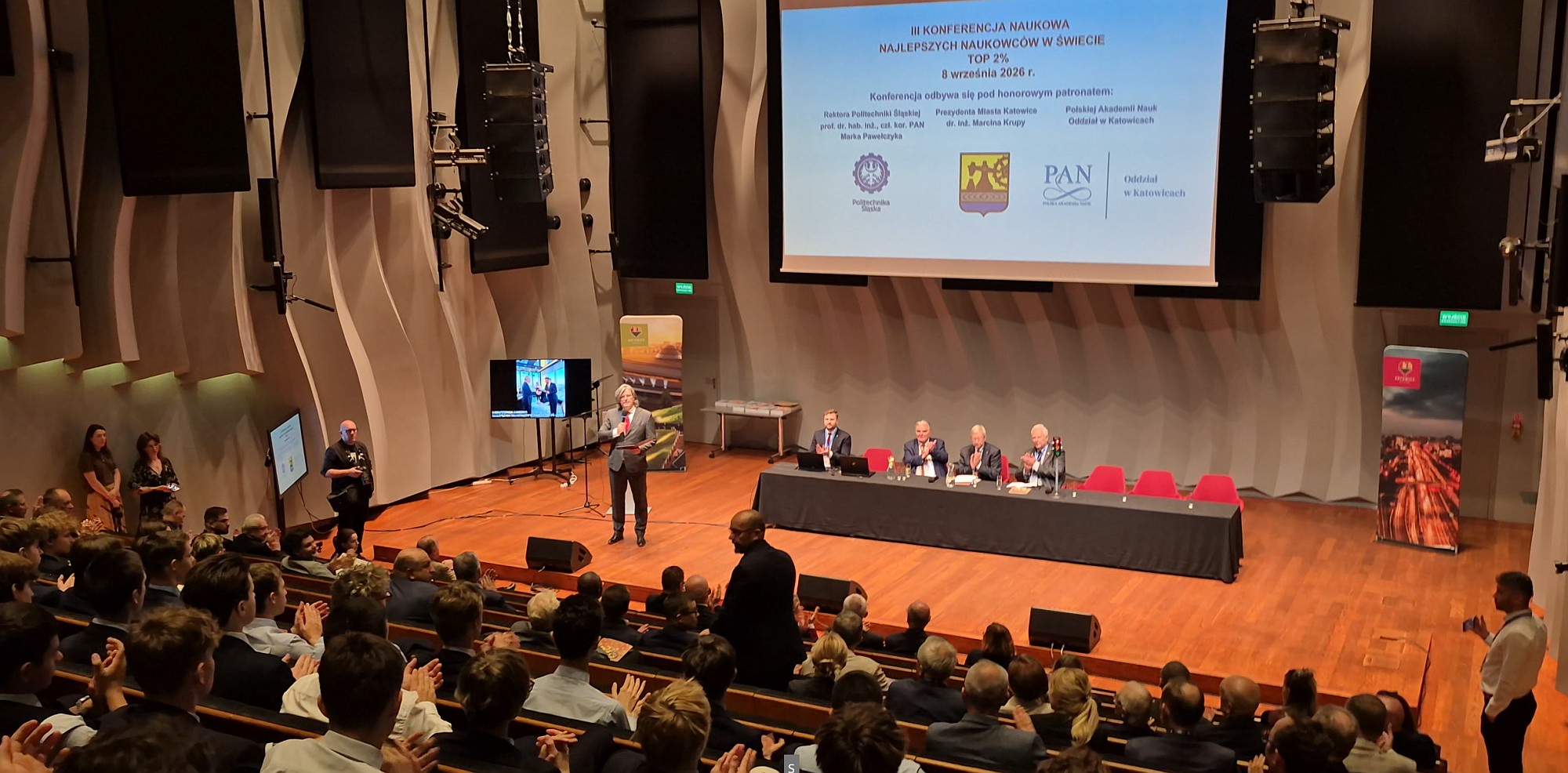 Konferencja Top 2% 2026 w NOSPR - przemawia przewodniczący komitetetu organizacyjnego, na wielkim ekranie plansza konferencji