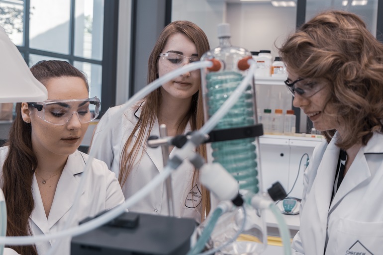 Na zdjęciu widzimy trzy studentki w białych fartuchach w laboratorium. Studentki stoją przy urządzeniach laboratoryjnych takich jak rurki czy pompki