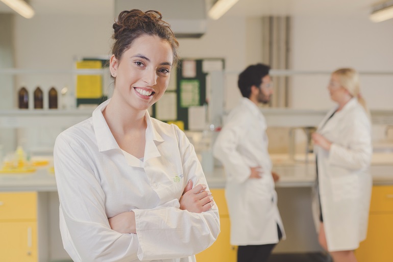 Zdjęcie studentów w laboratorium. Na pirwszym planie widzimy młoda Panią nuakowiec w tle kobieta i mężczyzna. wszyscy ubrani sa w białe fartuchy