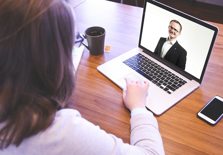 Na zdjęciu widzimy kobietę, która siedzi tyłem przy stole. Kobieta na długie brązowe włosy i biała bluzkę. Na stole stoi otwarty laptop. Na ekranie widzimy od pasa w grę mężczyznę w garniturze. Kobieta patrzy na ekran a rękę trzyma na klawiaturze. Obok laptopa stoi kubek