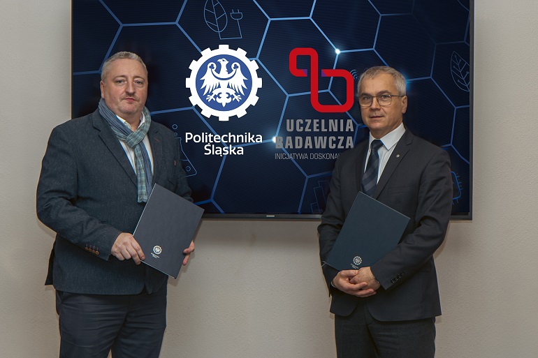 Na zdjęciu widzimy dwóch mężczyzn Rektora Politechniki oraz członka zarządu firmy Alstom. Panowie stoją za nimi ściana z dużą niebieską tablicą z logo Politechniki Śląskiej i Uczelni Badawczej