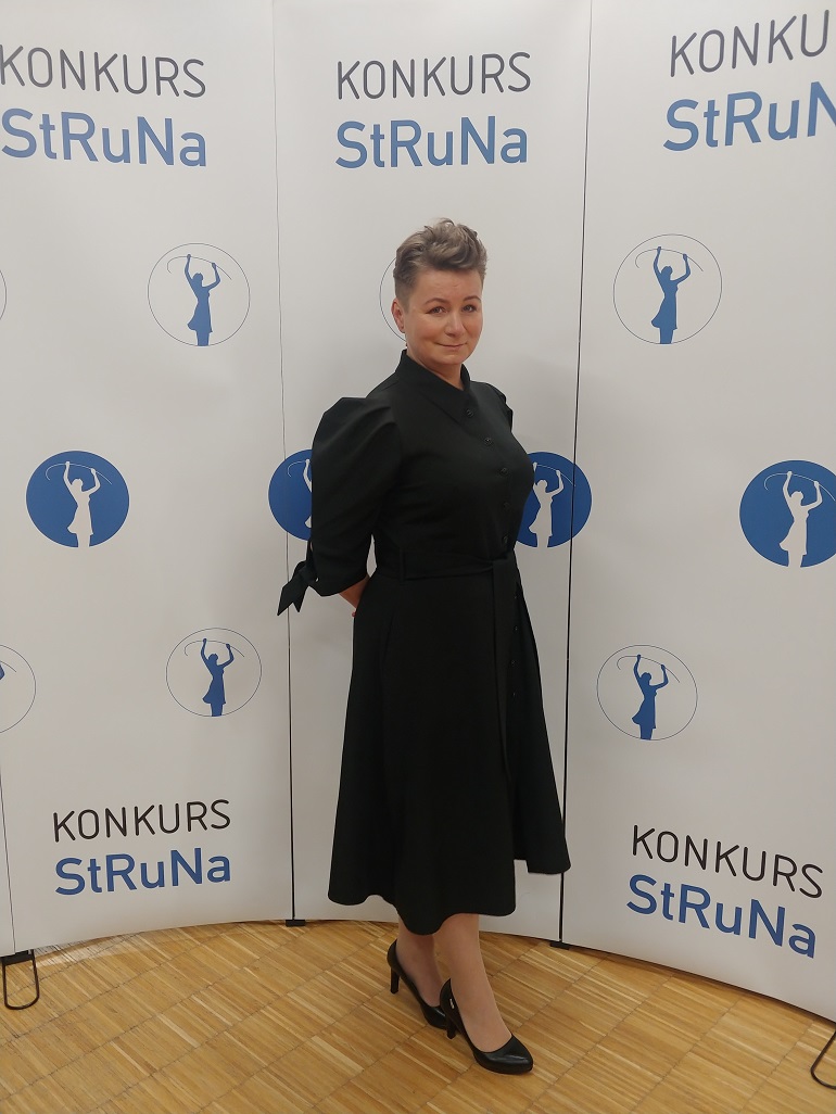 Na zdjęciu widzimy dr inż. Agatę Guzik Kopyto, która stoi w czarnej sukience. Za nią biała ściana z napisami Konkurs StRuNa