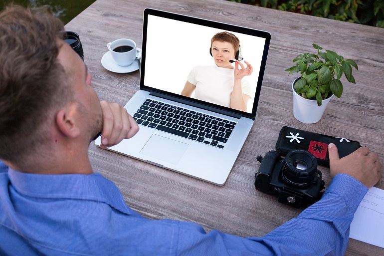 Na zdjęciu widzimy młodego mężczyznę siedzącego przed otwartym laptopem. Na ekranie monitora jest młoda kobieta w słuchawkach z mikrofonem. Na stole, na którym stoi laptop leży aparat fotograficzny, filiżanka z kawą oraz stoi kwiatek w białej doniczce