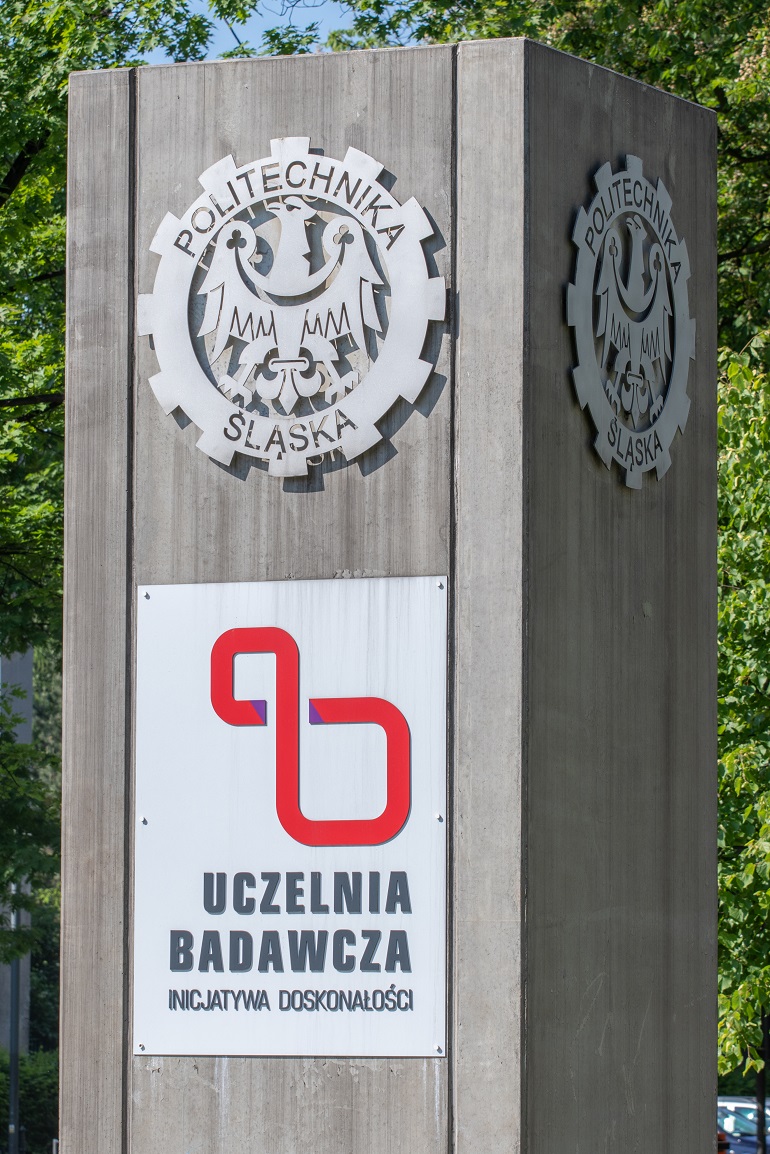 Na zdjęciu widzimy szary totem Politechniki Śląskiej. Na nim logo Uczelni oraz logo Uczelnia Badawcza