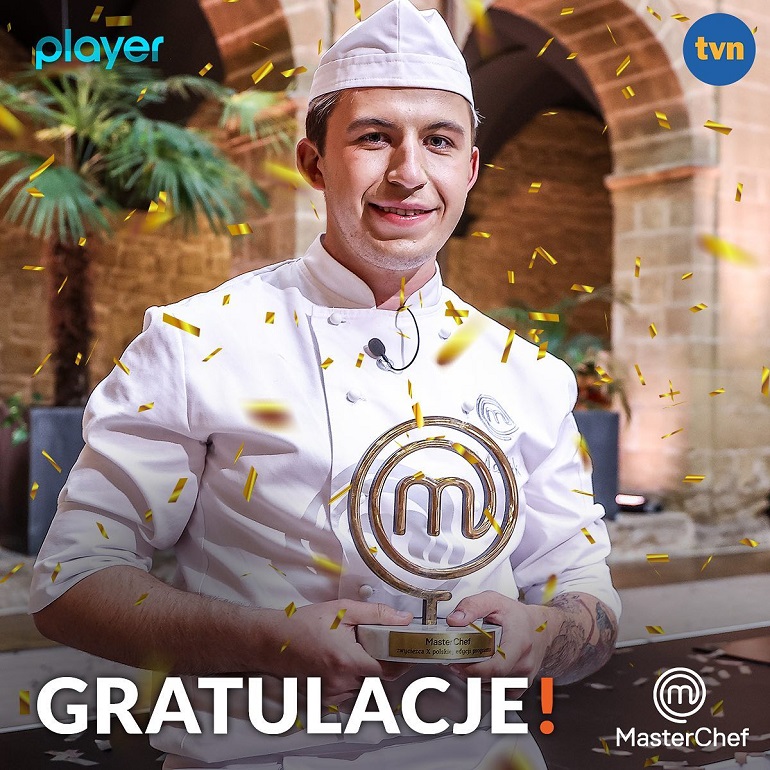 Na zdjęciu widzimy zwycięzcę 10 edycji konkursu master chef tvn. Na zdjęciu widzimy młodego chłopka w białym fartuchu i czapce kucharskiej trzymającego nagrodę konkursu. Na dole zdjęcie jest napisane gratulujemy