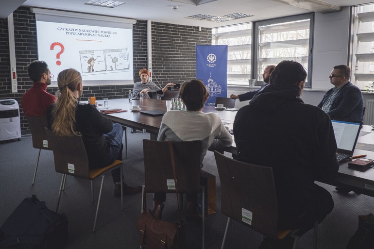 Na zdjęciu widzimy salę konferencyjną i duży stół, wokół którego siedzą uczestnicy szkolenia. Za prowadzącą wisi ekran, na którym jest slajd-prezentacja