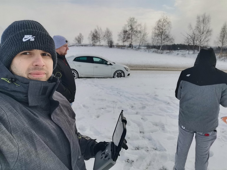 Na zdjęciu widzimy mężczyznę, który stoi w polu i robi sobie zdjęcie. W ręku trzyma mały komputerek. Na ziemi leży śnieg. W tle stoi białe auto oraz dwóch mężczyzn.