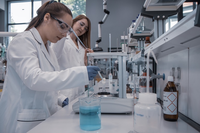 Na zdjęciu widzimy dwie studentki w białych fartuchach pracujące w laboratorium chemicznym