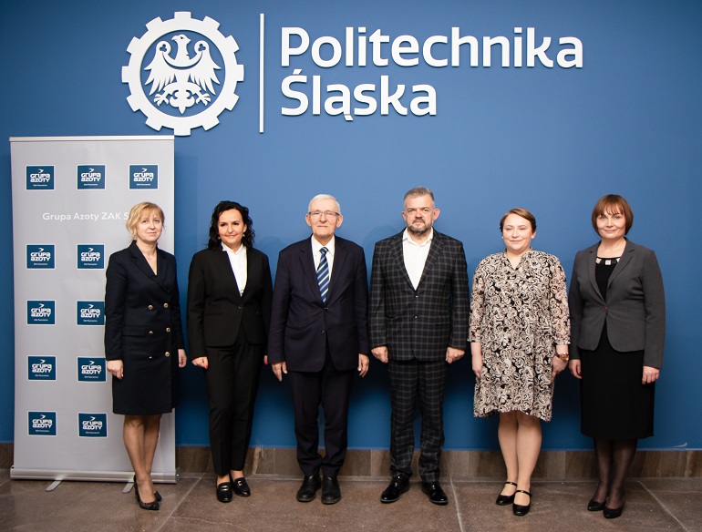 Na zdjęciu widzimy uczestników spotkania dotyczącego doktoratu wdrożeniowego, stojących na tle niebieskiej ściany z napisem Politechnika Śląska