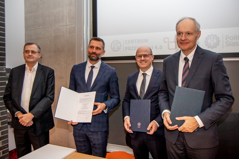 Na zdjęciu widzimy czterech mężczyznę, którzy stoją w Sali konferencyjnej. Trzech z nich trzyma teczki. Za nimi jest biały ekran