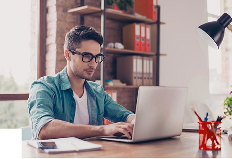 Na zdjęciu widzimy młodego chłopaka siedzącego przy laptopie. W tle regał z segregatorami