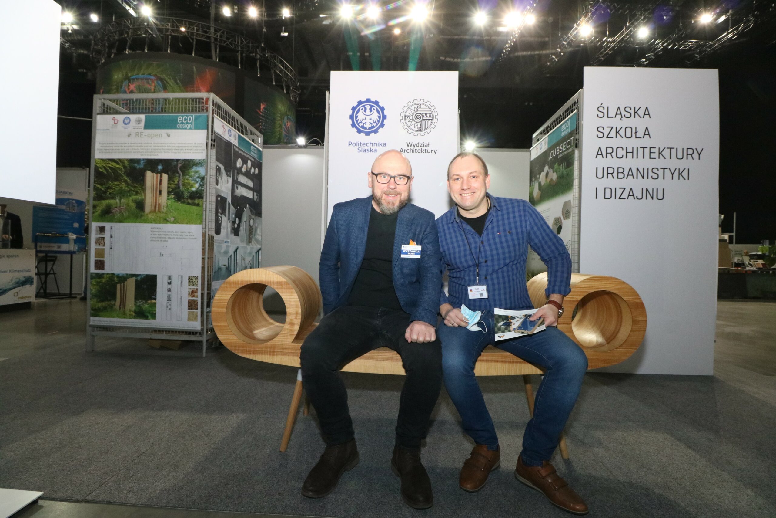Na zdjęciu widzimy dwóch mężczyzn siedzących na drewnianej ławce. Za nimi w tle stoisko targowe Wydziału Architektury