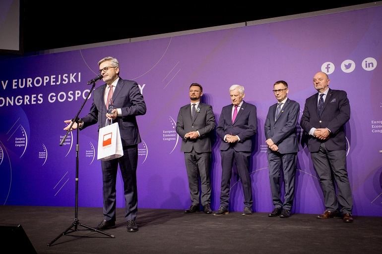 Na zdjęciu widzimy prezesa Tomasika strojącego na scenie przy mikrofonie za nim stoja czterej mężczyźni