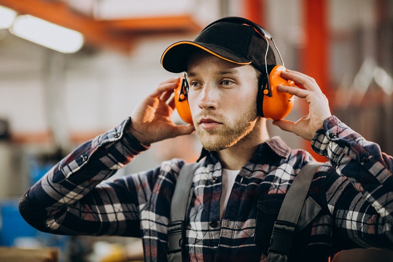 Na zdjęciu widzimy młodego mężczyznę w stroju roboczym. Na głowie ma czapkę z daszkiem. Ma założone robocze słuchawki, na których trzyma swoje ręce.