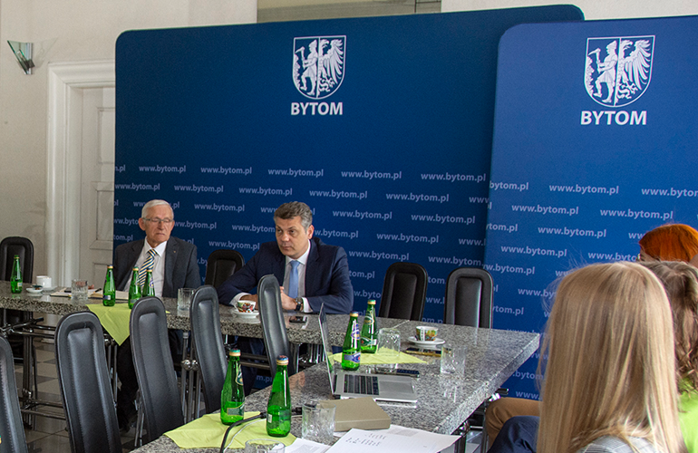 Zdjęcie przedstawia paru ludzi siedzących przy stole, w jakieś sali konferencyjnej.