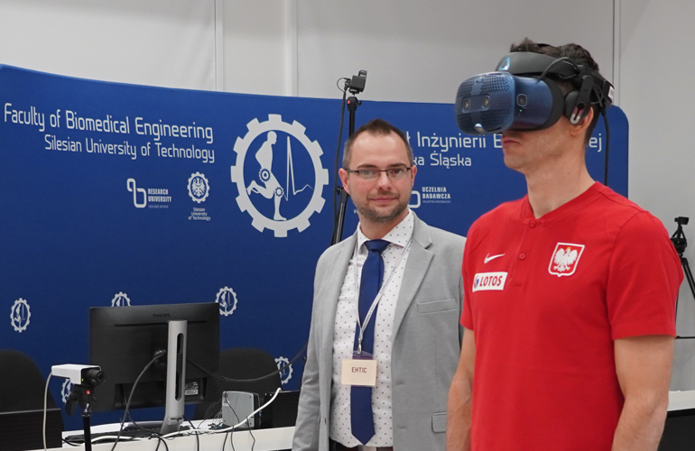 Zdjęcia przedstawia mężczyznę w czerwonej koszulce z goglami VR na twarzy, za nim stoi drugi mężczyzna.